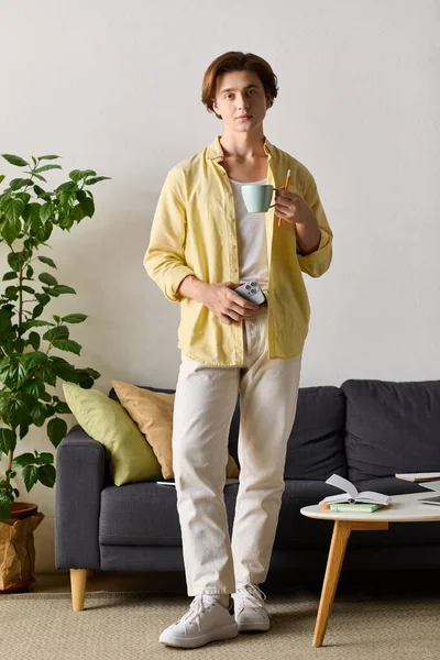 Ein stilvoller junger Mann steht in einem hellen Wohnzimmer, hält eine Tasse in der Hand und lächelt während einer Kaffeepause.. — Stockfoto