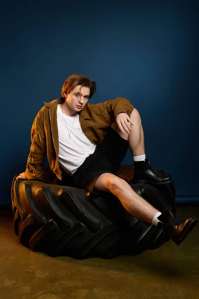 Handsome young man showcases stylish outfit while relaxing on a massive tire against a blue backdrop — Stock Photo