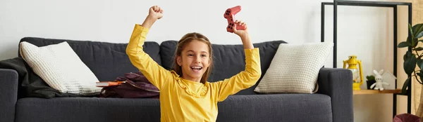 Ein junges Mädchen in leuchtend gelbem Outfit spielt zu Hause Videospiele und feiert fröhlich ihren Erfolg, Banner — Stockfoto