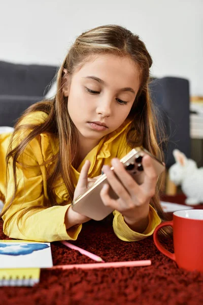 Una chica alfa gen vestida casualmente explora su teléfono inteligente mientras se relaja en la alfombra en casa. - foto de stock