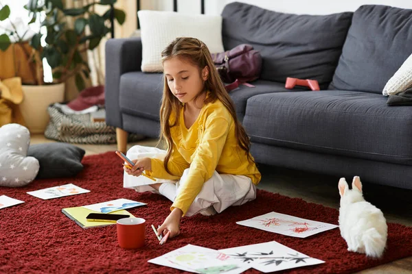 Ein Gen Alpha Mädchen sitzt auf dem Boden, umgeben von ihrem Kunstwerk und ihrem verspielten Hasen, verloren in Kreativität. — Stockfoto