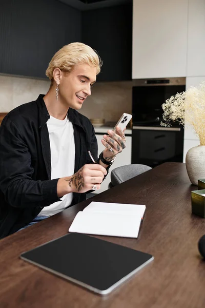 Schöner junger Mann sitzt mit seinem Smartphone an einem stilvollen Tisch, umgeben von einer gemütlichen Atmosphäre. — Stockfoto