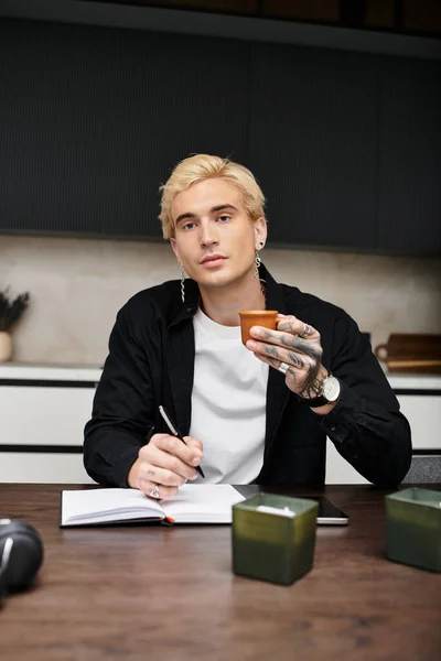 Ein junger Mann schreibt Gedanken in ein Notizbuch und nippt an einem Drink in seiner schicken Wohnung. — Stockfoto