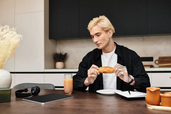 Привлекательный молодой человек сидит за кухонным столом, наслаждаясь круассаном, потягивая сок. — стоковое фото