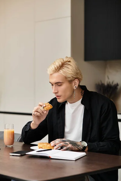 Um jovem bonito saboreia um doce em sua mesa de jantar, imerso em um bom livro. — Fotografia de Stock
