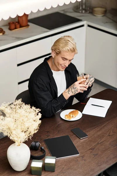 Ein junger Mann mit Tätowierungen lächelt, während er ein Croissant genießt und in einer modernen Küche an einem Laptop arbeitet. — Stockfoto
