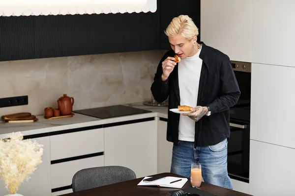 Красивый молодой человек кусается в выпечку, стоя на стильной кухне, чувствуя себя довольным. — стоковое фото