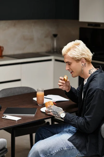 Um jovem morde uma pastelaria em uma mesa moderna enquanto usa seu laptop. — Fotografia de Stock