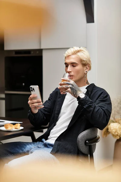 A young handsome man relaxes in a modern apartment, sipping a drink and checking his phone. — Stock Photo
