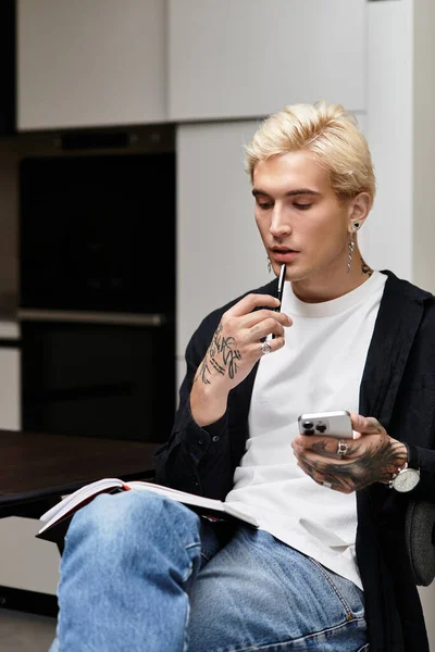 Un joven reflexivo se sienta en un apartamento elegante, sosteniendo una pluma y un teléfono. - foto de stock
