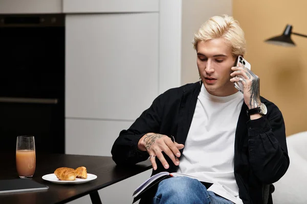 Un joven se sienta en una mesa, conversando casualmente por teléfono mientras desayunaba. - foto de stock
