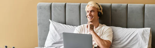 A young man in a white shirt sits on his bed, smiling and enjoying music with headphones on. — Stock Photo