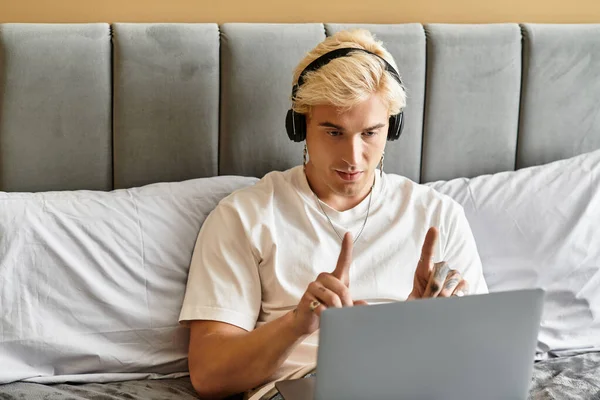 Молодой человек в наушниках сидит на кровати и занимается виртуальной встречей. — стоковое фото