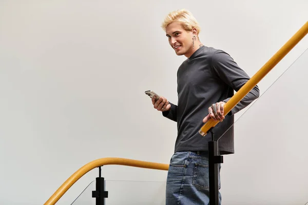 Handsome young man stands on a staircase, smiling while using his phone in a modern apartment. — Stock Photo