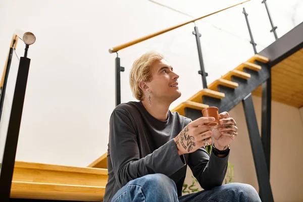 A young handsome man with tattoos sits comfortably, savoring a warm drink in his modern apartment. — Stock Photo