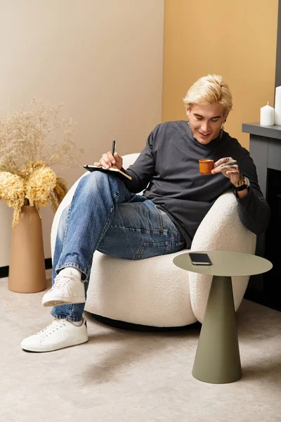 En casa, un joven saborea su café, tomando notas mientras se relaja en una silla elegante. - foto de stock