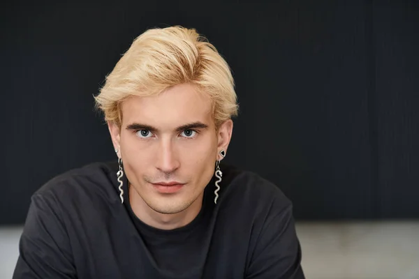 Молодой, красивый мужчина с светлыми волосами и уникальными серьгами уверенно смотрит в стильную квартиру. — стоковое фото