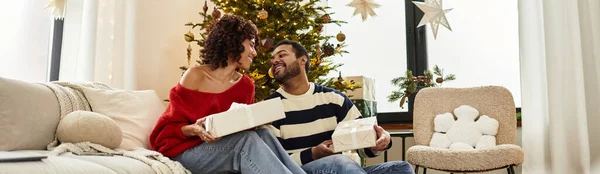 Щаслива пара ділиться сміхом і обміном подарунками за їх красиво прикрашеною ялинкою, банер — стокове фото
