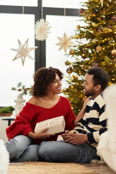 Ein freudiges afroamerikanisches Paar teilt einen besonderen Weihnachtsmoment am geschmückten Baum. — Stockfoto