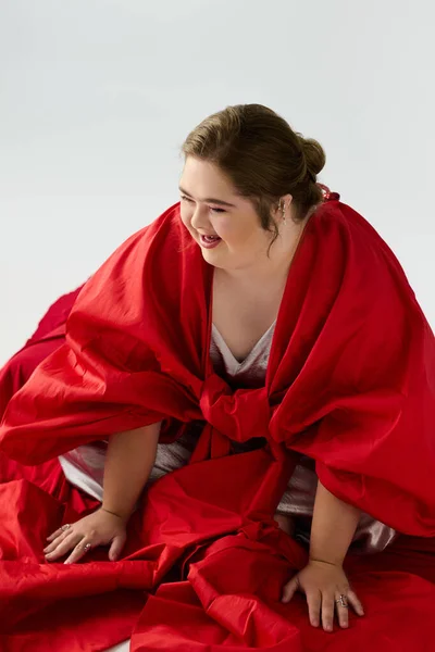 A joyful young woman with Down syndrome smiles amid flowing red fabric, highlighting her spirit. — Stock Photo