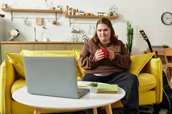Eine junge Frau mit Down-Syndrom sitzt gemütlich zu Hause auf einem gelben Sofa und nippt am Kaffee. — Stockfoto
