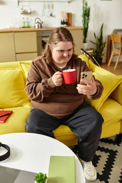 Young woman with Down syndrome sits on a yellow couch, looking at her phone with a red cup. — Stock Photo