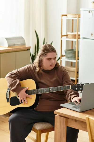 Молода жінка з синдромом Дауна сидить за столом, граючи на гітарі, фокусуючись на своєму ноутбуці. — стокове фото