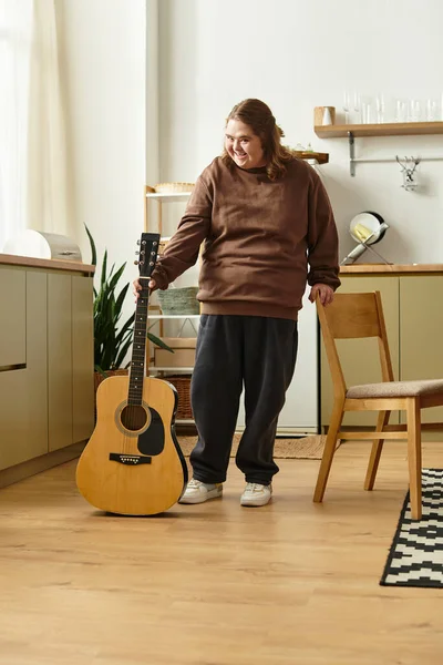 Une jeune femme trisomique se tient joyeusement à côté d'une guitare dans sa maison confortable, prête à jouer. — Photo de stock