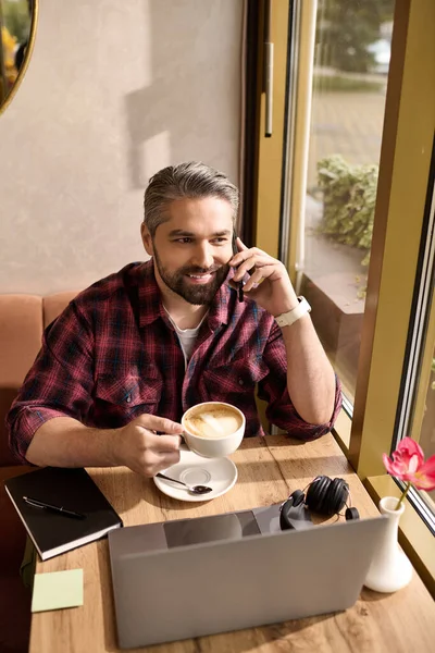 A handsome, mature man dressed stylishly sips coffee and chats happily on the phone indoors. — Stockfoto