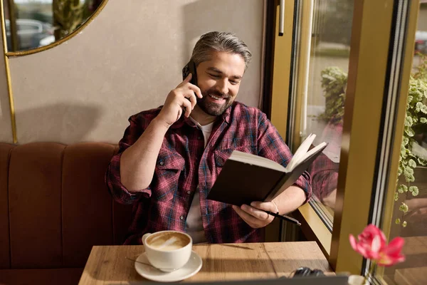 A handsome mature man reads a book and talks on the phone, sipping coffee in a warm cafe. — Stockfoto