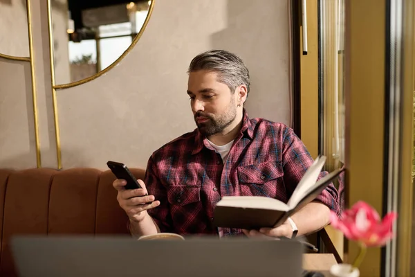 Handsome man in fashionable attire sits in a cozy cafe, reading a book and checking his phone. — Stockfoto
