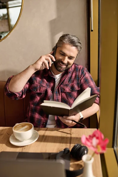 A stylish man chats happily on the phone, reading a book and savoring coffee in a cozy location. — Stockfoto