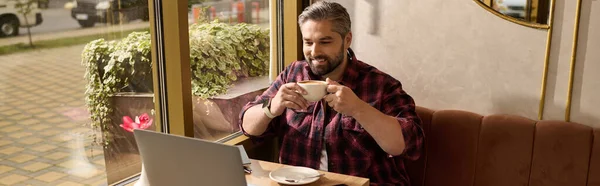A charming man in stylish attire happily sips coffee while engaging with his laptop in a cozy cafe. — Stockfoto