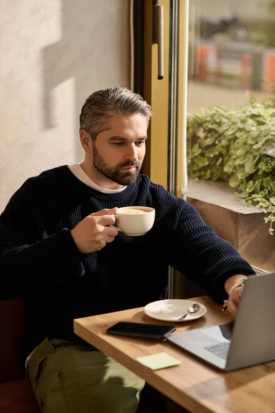 Handsome man in stylish attire savors his coffee and focuses on his laptop in a cozy cafe. — Stockfoto
