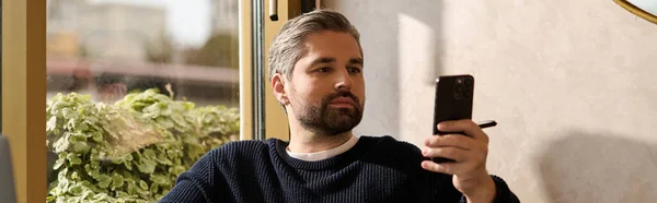 A handsome, mature man relaxes by the window, focused on his smartphone screen with calm demeanor. — Stockfoto