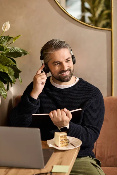 Handsome man in stylish attire smiles while listening to music with a notebook and dessert nearby. — Stockfoto