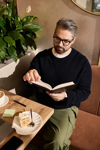 Sitting in a modern cafe, a mature man is engrossed in a book while savoring dessert. — Stockfoto