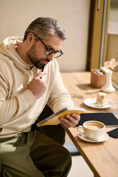 A mature man with silver hair sits in a trendy cafe, savoring coffee and jotting down thoughts. — Stockfoto