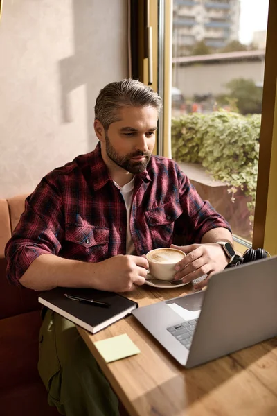 Handsome man in stylish attire relaxes with coffee, focused on his laptop during daytime. — Stockfoto