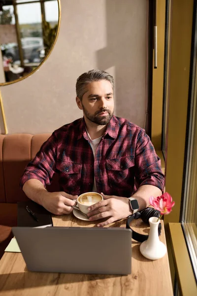 A mature man in stylish attire sips his coffee while focused on his laptop in a cafe. — Stockfoto