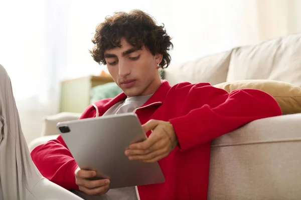 Handsome man dressed in trendy attire sits on sofa, focused on his tablet in a bright room. — Stock Photo