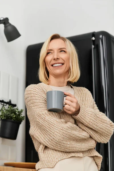 A blonde woman dressed in cozy attire smiles while holding a cup in her stylish home. — Stock Photo