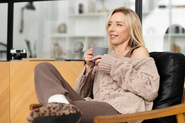 Eine blonde Frau entspannt sich in ihrer stilvollen Wohnung, nippt an einem warmen Getränk und umarmt den Komfort. — Stockfoto