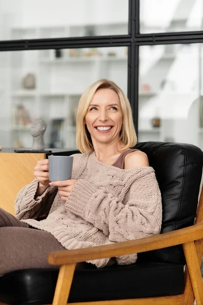 Блондинка в зручному одязі відпочиває з теплим напоєм у своєму сучасному будинку. — стокове фото