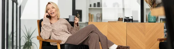 Una mujer rubia se relaja en casa con ropa cómoda, sorbiendo té y charlando por teléfono, pancarta - foto de stock