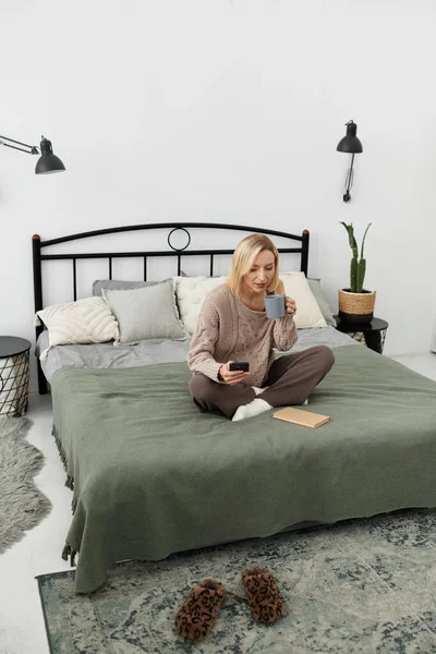 Eine blonde Frau sitzt gemütlich auf ihrem Bett, nippt an Tee und entspannt sich mit einem Buch. — Stockfoto