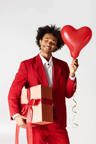 Handsome young man smiles while holding a gift box and a heart shaped balloon with joy. — Stockfoto