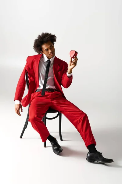 Young man in a sharp red suit sits confidently on a chair, holding a heart shaped box with flair. — Stockfoto