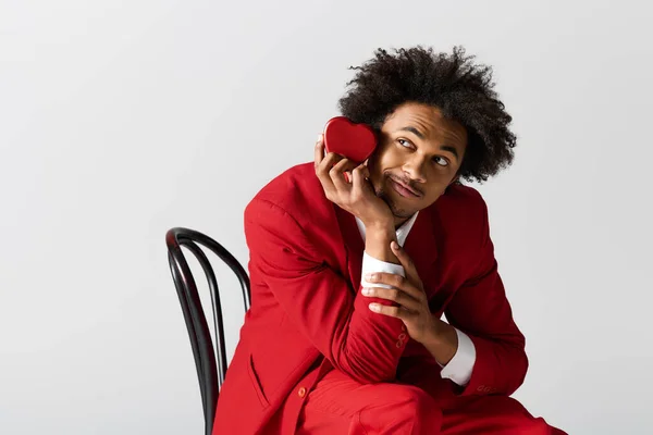 Handsome young man in stylish attire sits on a chair, holding a heart shaped token with joy. - foto de stock