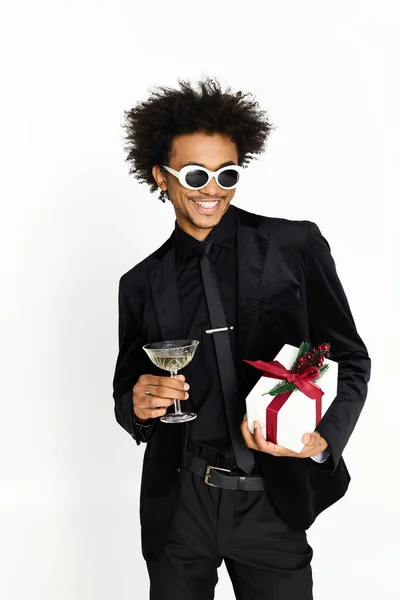 Handsome young man in black attire holds a cocktail and a present, radiating happiness. — Stock Photo